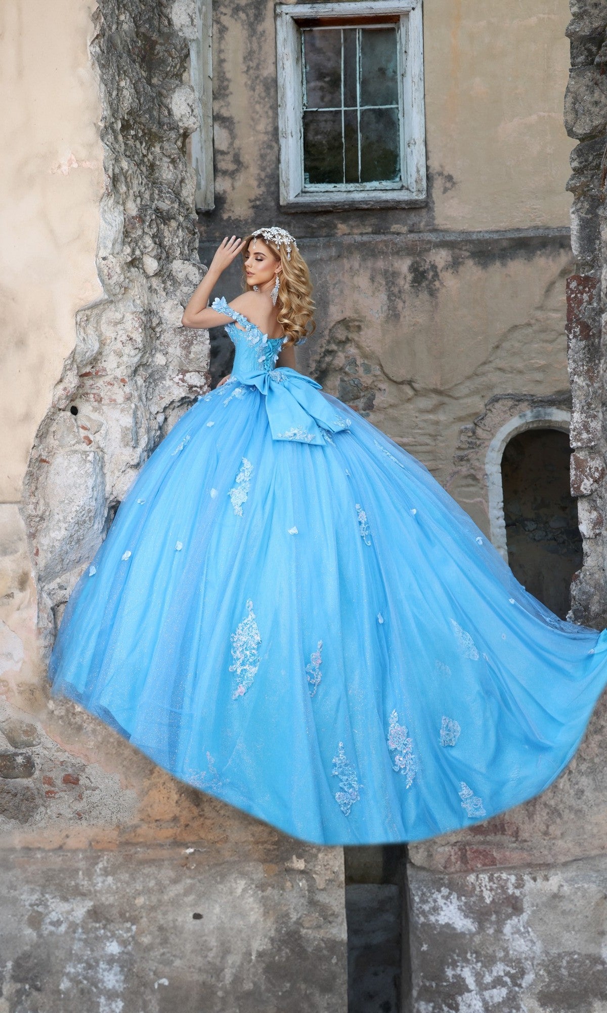 A woman in an off-the-shoulder Tiffany-blue Quinceanera Dress Q120 by Nina Canacci stands against an old, weathered stone wall with a window.